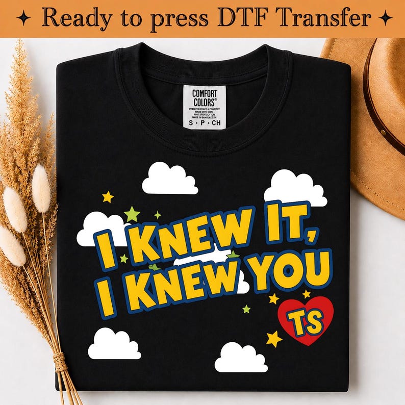 May include: A black t-shirt featuring a graphic print with white clouds, yellow stars, and the text "I KNEW IT, I KNEW YOU" in yellow with a blue outline. A red heart contains the yellow letters "TS". The shirt has a Comfort Colors brand label.