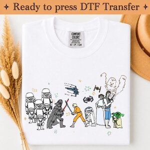 Könnte beinhalten: Ein weißes T-Shirt mit einer bunten, handgezeichneten Grafik von Star Wars-Figuren, darunter Stormtrooper, Darth Vader, Luke Skywalker, R2-D2, Prinzessin Leia, Han Solo, Chewbacca und Yoda, sowie verschiedenen Raumschiffen und Sternen.