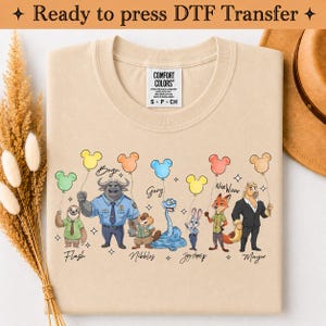 Puede incluir: Una camiseta beige con un estampado gr&aacute;fico de siete personajes de dibujos animados de Zootopia, cada uno sosteniendo un globo de color con forma de cabeza de rat&oacute;n. Los personajes est&aacute;n etiquetados con nombres como Flash, Bogo, Nibbles, Gary, Judy Hopps, Nick Wilde y Mayor.
