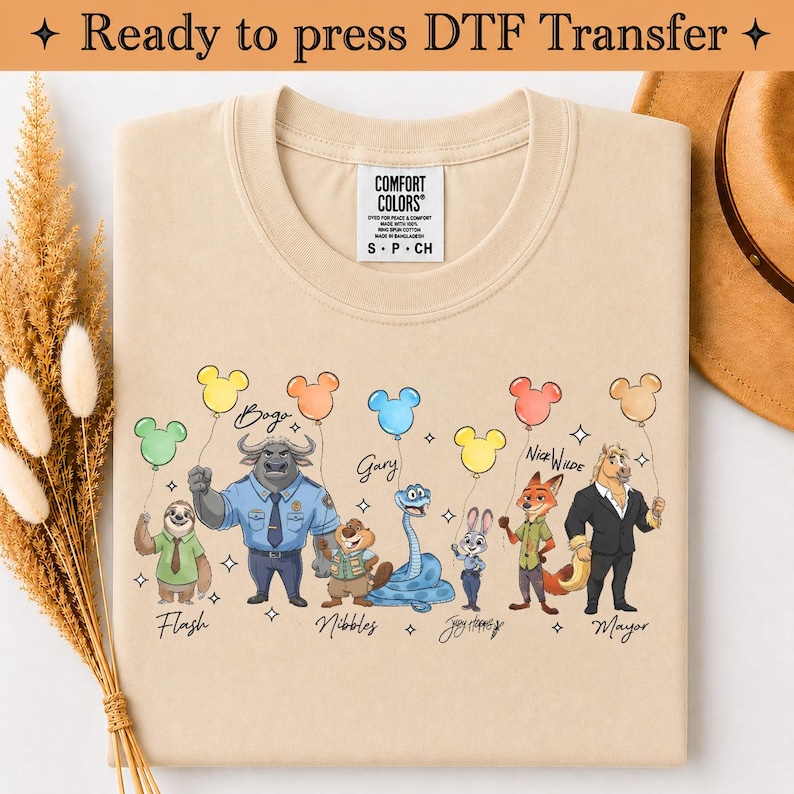 Puede incluir: Una camiseta beige con un estampado gr&aacute;fico de siete personajes de dibujos animados de Zootopia, cada uno sosteniendo un globo de color con forma de cabeza de rat&oacute;n. Los personajes est&aacute;n etiquetados con nombres como Flash, Bogo, Nibbles, Gary, Judy Hopps, Nick Wilde y Mayor.