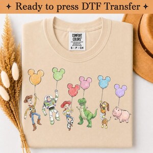 Puede incluir: Una camiseta beige con un estampado gr&aacute;fico de seis personajes animados sosteniendo globos de colores con forma de Mickey Mouse. Los personajes incluyen un vaquero, un guardi&aacute;n espacial, una vaquera, un dinosaurio verde, un perro y un cerdo.