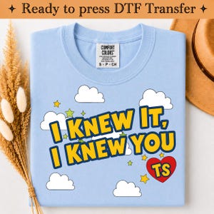 May include: A light blue t-shirt featuring a graphic print with white clouds, yellow stars, and a red heart containing the letters TS. The text reads I KNEW IT, I KNEW YOU in bold yellow letters with a dark blue outline. The shirt has a Comfort Colors label.