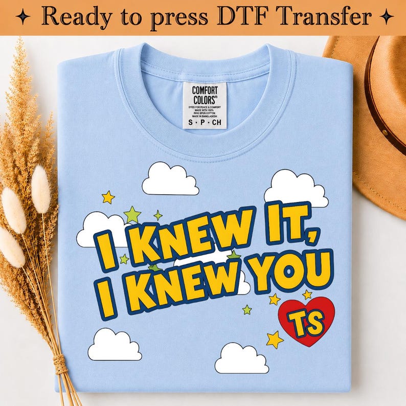 May include: A light blue t-shirt featuring a graphic print with white clouds, yellow stars, and a red heart containing the letters TS. The text reads I KNEW IT, I KNEW YOU in bold yellow letters with a dark blue outline. The shirt has a Comfort Colors label.