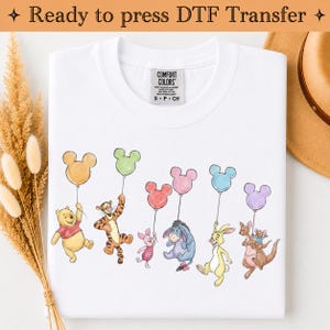 May include: A white t-shirt featuring a graphic print of Winnie the Pooh, Tigger, Piglet, Eeyore, Rabbit, Kanga, and Roo, each holding a Mickey Mouse-shaped balloon in various colors. The shirt has a Comfort Colors brand label at the neck.