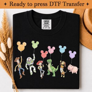 Puede incluir: Una camiseta negra con un estampado gr&aacute;fico de seis personajes animados, cada uno sosteniendo un globo con forma de Mickey Mouse en varios colores pastel. La camiseta tiene una etiqueta interior blanca con el texto Comfort Colors S P CH.