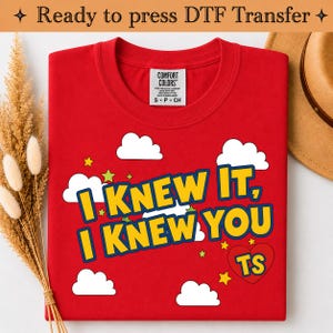 May include: A red t-shirt featuring the text I KNEW IT, I KNEW YOU TS in yellow letters with blue outlines. The design includes white clouds and small yellow and green stars, with the letters TS inside a red heart. The shirt has a Comfort Colors label.
