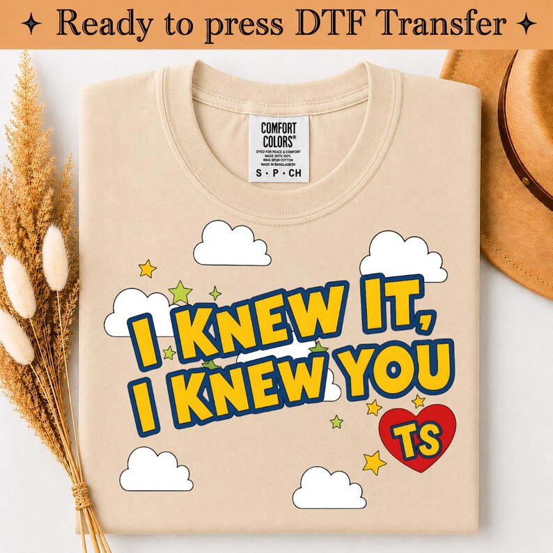 May include: A beige t-shirt featuring a graphic print with white clouds, small yellow and green stars, and a red heart containing the letters TS. The text reads I KNEW IT, I KNEW YOU in bold yellow letters with a dark blue outline.