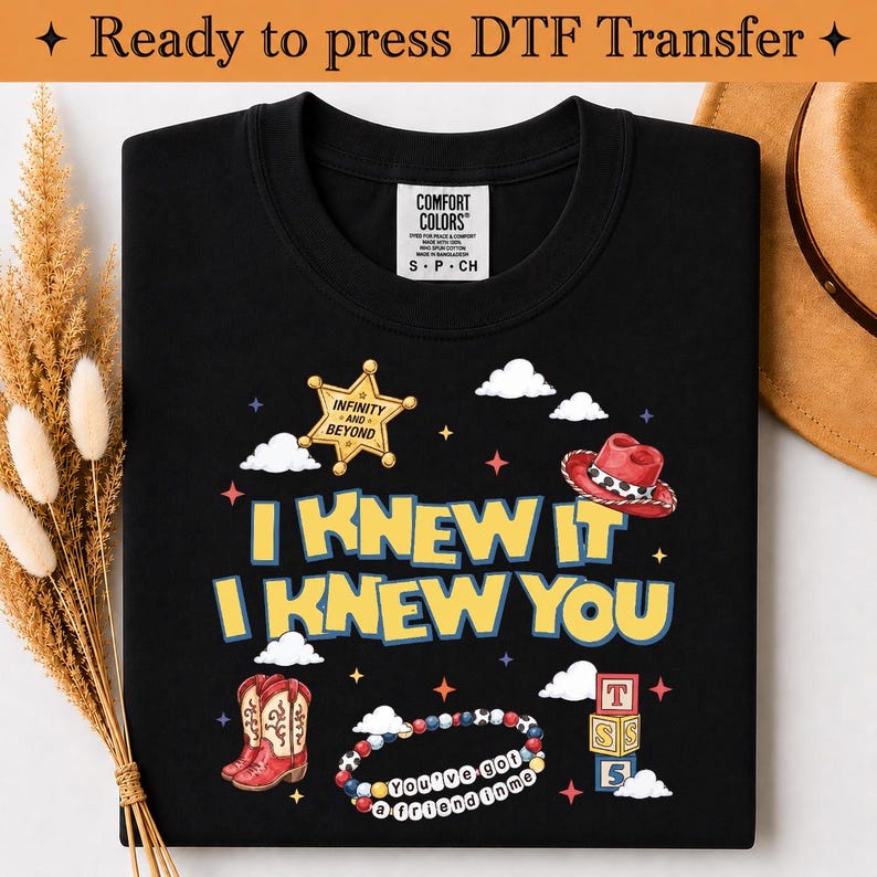 May include: A black t-shirt featuring a graphic print with the text I KNEW IT I KNEW YOU, a yellow sheriff star with the text INFINITY AND BEYOND, a red cowboy hat, red cowboy boots, a beaded bracelet with the text YOU'VE GOT A FRIEND IN ME, and blocks.