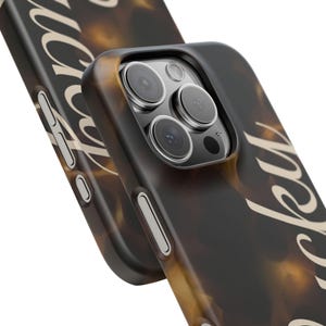 Può includere: Primo piano di due custodie per telefono con un motivo a scaglie di tartaruga nei toni del marrone e beige. Le custodie hanno un bordo scuro e la scritta "lucky" è visibile in corsivo. Le lenti della fotocamera del telefono sono visibili.
