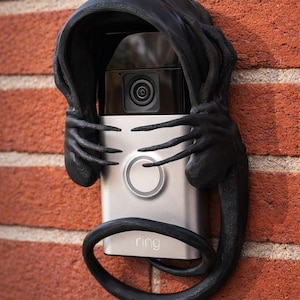 Puede incluir: Una cubierta decorativa negra para un timbre de video Ring, montada en una pared de ladrillos rojos. La cubierta presenta un diseño de criatura con manos con garras y una cola curvada, agregando una estética única a la seguridad del hogar.