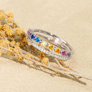 May include: A silver ring with a rainbow of gemstones and a row of small white stones. The ring is set on a beige surface with a sprig of yellow flowers.