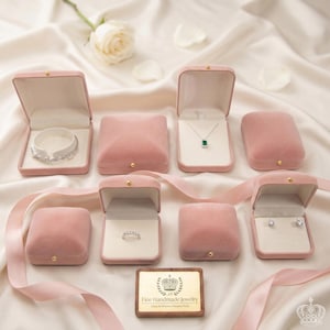 May include: An assortment of pink velvet jewelry boxes, some open to display a bracelet, necklace, ring, and earrings. A wooden plaque with gold lettering reads "Fine Handmade Jewelry". A white rose and ribbon are in the background.
