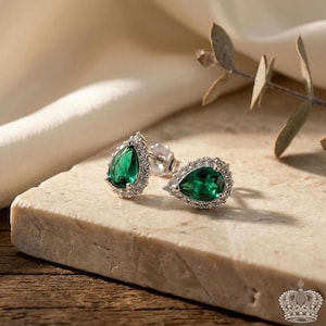 May include: A pair of stud earrings featuring pear-shaped, emerald-green gemstones surrounded by a halo of small, clear stones. The earrings are set in a silver-colored metal and rest on a light-colored stone surface.