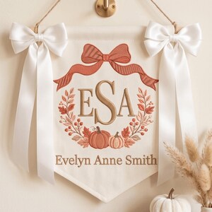 May include: A cream-colored fabric wall banner featuring an embroidered rust-colored bow at the top, a central monogram with the letters ESA, and a wreath of autumn leaves and pumpkins. The name Evelyn Anne Smith is embroidered below.