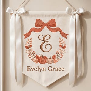May include: A cream-colored fabric wall banner featuring an embroidered rust-colored bow at the top, a central letter E, and a wreath of autumn leaves and pumpkins. The name Evelyn Grace is embroidered below. White satin ribbons hang from the top.
