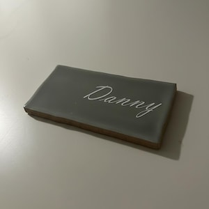 May include: A rectangular, grey tile with the name "Danny" written in elegant white script. The tile has a smooth, glossy surface and a light brown edge, resting on a white surface.