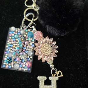 May include: A decorative keychain featuring a black fluffy pom-pom, a rectangular charm covered in colorful rhinestones, a pink rhinestone sunflower, and a silver letter "H" encrusted with rhinestones. A small heart charm hangs below the letter.