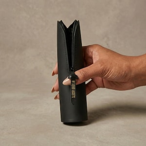 May include: A black cylindrical cosmetic bag with a zipper closure. The bag is held open, revealing the interior. The zipper pull has the text "MY LIQUID ASSETS" printed on it. The bag is made of a matte material.