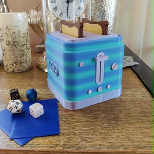 Puede incluir: Una tostadora a rayas azules y verdes con dos rebanadas de pan tostado. La tostadora tiene una palanca y botones. Dados y un cuadrado azul están en primer plano. Un reloj y una vela están en el fondo.