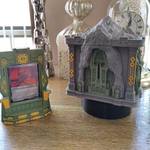 Puede incluir: Un soporte de plástico verde y dorado para tarjetas que sostiene una carta coleccionable Witch-king of Angmar, junto a una estructura de fortaleza de montaña impresa en 3D gris y verde con detalles arquitectónicos y acentos dorados.