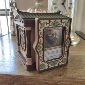 May include: A brown and cream-colored deck box featuring a green roof and ornate floral detailing. The front panel displays a Lathril, Blade of the Elves trading card within a decorative frame. The structure includes columns and a side emblem.
