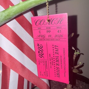 May include: A rectangular pink tag with black text resembling a concert ticket. It reads COACH, LIVE YOUR STORY, ON TOUR, NEW YORK, NEW YORK, AUG. 18, 2026, SEC C, ROW PP, SEAT 41. The tag hangs from a gold-colored ball chain.