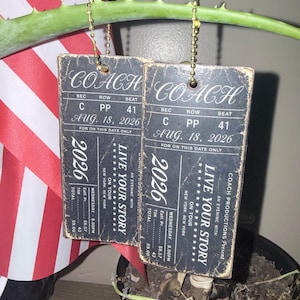 May include: Two rectangular black tags with distressed edges, each featuring white text resembling a concert ticket. The text reads COACH, SEC C, ROW PP, SEAT 41, AUG. 18, 2026, and LIVE YOUR STORY. Each tag hangs from a gold-colored metal ball chain.