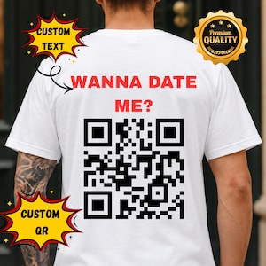 Puede incluir: Una camiseta blanca con el texto rojo WANNA DATE ME? sobre un gran código QR en blanco y negro. La imagen incluye gráficos de estrellas amarillas con el texto CUSTOM TEXT y CUSTOM QR, además de un sello dorado Premium Quality en la esquina.