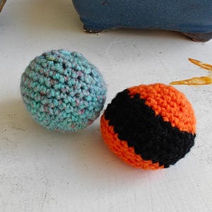 May include: Two spherical crochet balls. One ball features a variegated light blue and teal yarn with subtle flecks of color. The second ball is crafted with alternating horizontal bands of bright orange and solid black yarn in a textured stitch.
