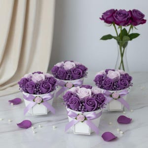 以下が含まれることがあります： 紫と薄ピンクのバラが詰められた4つの白い容器。ラベンダー色のリボンと小さなバラのアクセントで結ばれています。紫色のバラの花瓶が背景にあり、花びらと真珠が散らばっています。