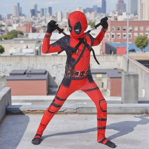 Op de afbeelding: Een rood en zwart Deadpool-kostuum met een masker, handschoenen en twee speelgoedzwaarden. Het kostuum heeft een gereedschapsriem en zwarte accenten. De persoon staat op een dak met een skyline op de achtergrond.