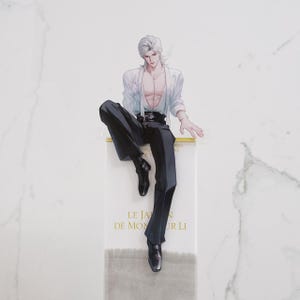 May include: A rectangular bookmark featuring an illustration of a person with silver hair, wearing a white open-front shirt and black trousers. The lower portion of the bookmark is white with gold text reading LE JARDIN DE MONSIEUR LI.