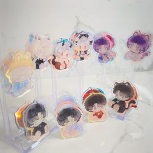 May include: A collection of holographic acrylic charms featuring cartoon characters in various outfits. The charms have a clear, iridescent finish and are displayed on a clear plastic stand. The characters have different hair colors and wear outfits like frog hats and cat ears.