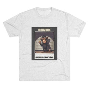 Puede incluir: Camiseta gris claro con un gráfico de un mono en una foto policial. El mono sostiene un plátano. La palabra "DRUNK" está encima de la imagen. Debajo de la imagen, el texto dice "CHARGED WITH PUBLIC INTOXICATION FIGHTING WITH DRUNK RACCOON."