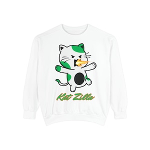 May include: White sweatshirt featuring a cartoon cat with green and black accents, spewing fire. The text "Kat Zilla" is printed in green and gold below the cat illustration. The sweatshirt has long sleeves and a crew neck.