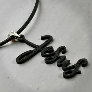 May include: Black pendant necklace with the word "Jesus" in a script font. The pendant is attached to a black cord with a silver-colored clasp. The necklace is displayed on a light gray surface.