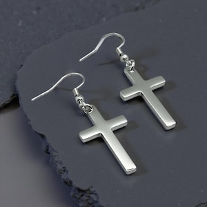 May include: A pair of silver-colored cross earrings. Each earring features a simple cross pendant suspended from a curved ear wire. The earrings are displayed on a dark gray, textured surface.