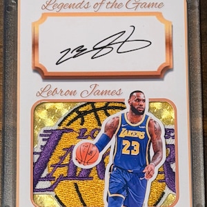 May include: A collectible sports trading card featuring a black ink autograph of Lebron James above a patch of a Lakers jersey. The card displays the text Legends of the Game, Lebron James, and One of One, encased in a clear protective plastic holder.