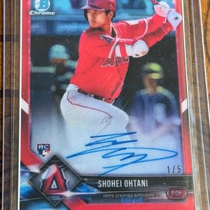 以下が含まれることがあります： 保護用プラスチックケースに入った大谷翔平のBowman Chromeルーキー野球カード。赤い枠、青いインクのサイン、'1/5'、'RC'、'P/DH'の文字が特徴です。選手はロサンゼルス・エンゼルスの赤いユニフォームを着てバットを振る姿が描かれています。