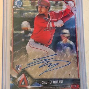 May include: A Bowman Chrome Shohei Ohtani baseball card in a protective plastic case. The card features a holographic background, a red Angels uniform, a blue ink autograph, a rookie card logo, and the text 1/1, Shohei Ohtani, and Topps Certified.