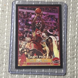 May include: A basketball trading card featuring LeBron James in a Cleveland Cavaliers uniform. The card has a red and black border with the text "DRAFT PICK #1" and "2003-2004". The image shows James shooting a basketball.