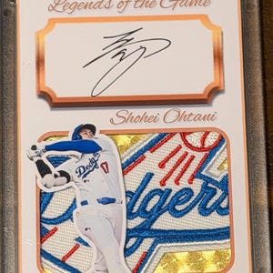 May include: A Legends of the Game trading card featuring an autograph by Shohei Ohtani. The card displays a die-cut image of the player in a white and blue Dodgers uniform swinging a bat, set against a gold-textured background with embroidery.
