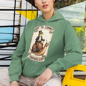 May include: Sage green hoodie featuring a humorous illustration. The graphic shows a frog in a top hat riding a snail, with the text "OFF TO COMMIT TOMFOOLERY". The hoodie has a drawstring hood and a front pocket.