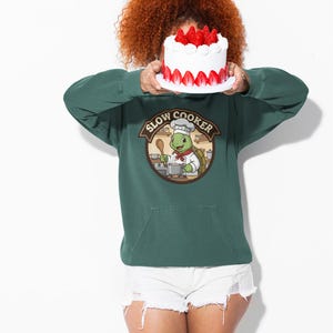 May include: A green hooded sweatshirt with a kangaroo pocket and a graphic of a turtle chef with the words "Slow Cooker". The turtle is wearing a chef's hat and holding a wooden spoon. A white cake with strawberries on top is held in front of the face.