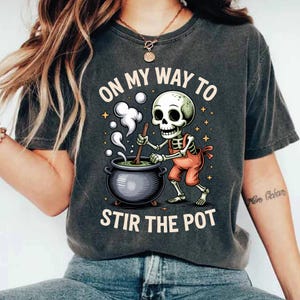 May include: Dark gray t-shirt with the text "ON MY WAY TO STIR THE POT". The graphic shows a skeleton in orange overalls stirring a cauldron with steam rising. The design includes stars and a cartoon style.