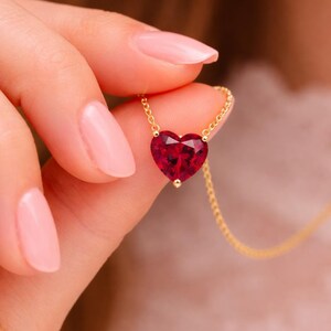 May include: A gold necklace featuring a heart-shaped, faceted, deep red gemstone pendant. The pendant is suspended from a delicate gold chain. The necklace is held up against a soft, blurred background.