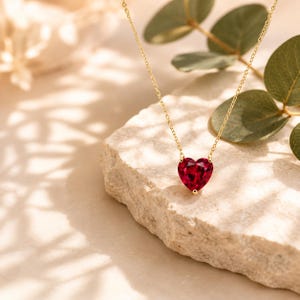 May include: A delicate gold chain necklace featuring a single, faceted, heart-shaped red gemstone pendant. The pendant is securely held by a small gold setting, and the thin chain is attached to both sides of the heart, creating a simple, elegant piece.