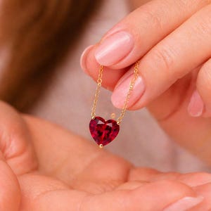 Puede incluir: Un collar con un colgante de piedra preciosa en forma de corazón de color rojo intenso y vibrante, suspendido de una cadena fina y delicada de tono dorado. La piedra facetada refleja la luz, mostrando su color saturado y diseño puntiagudo.