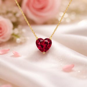 Pode incluir: Um colar de ouro com um pingente de pedra preciosa vermelha facetada em forma de coração. O pingente é suspenso por uma delicada corrente de ouro. O fundo apresenta rosas cor-de-rosa suaves e tecido de cetim branco.