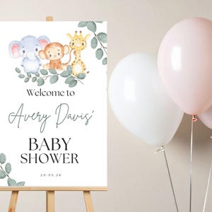 May include: A white rectangular sign on a wooden easel featuring watercolor illustrations of a blue elephant, a brown monkey, and a yellow giraffe surrounded by green eucalyptus leaves. The text reads Welcome to Avery Davis' BABY SHOWER 20.05.26.